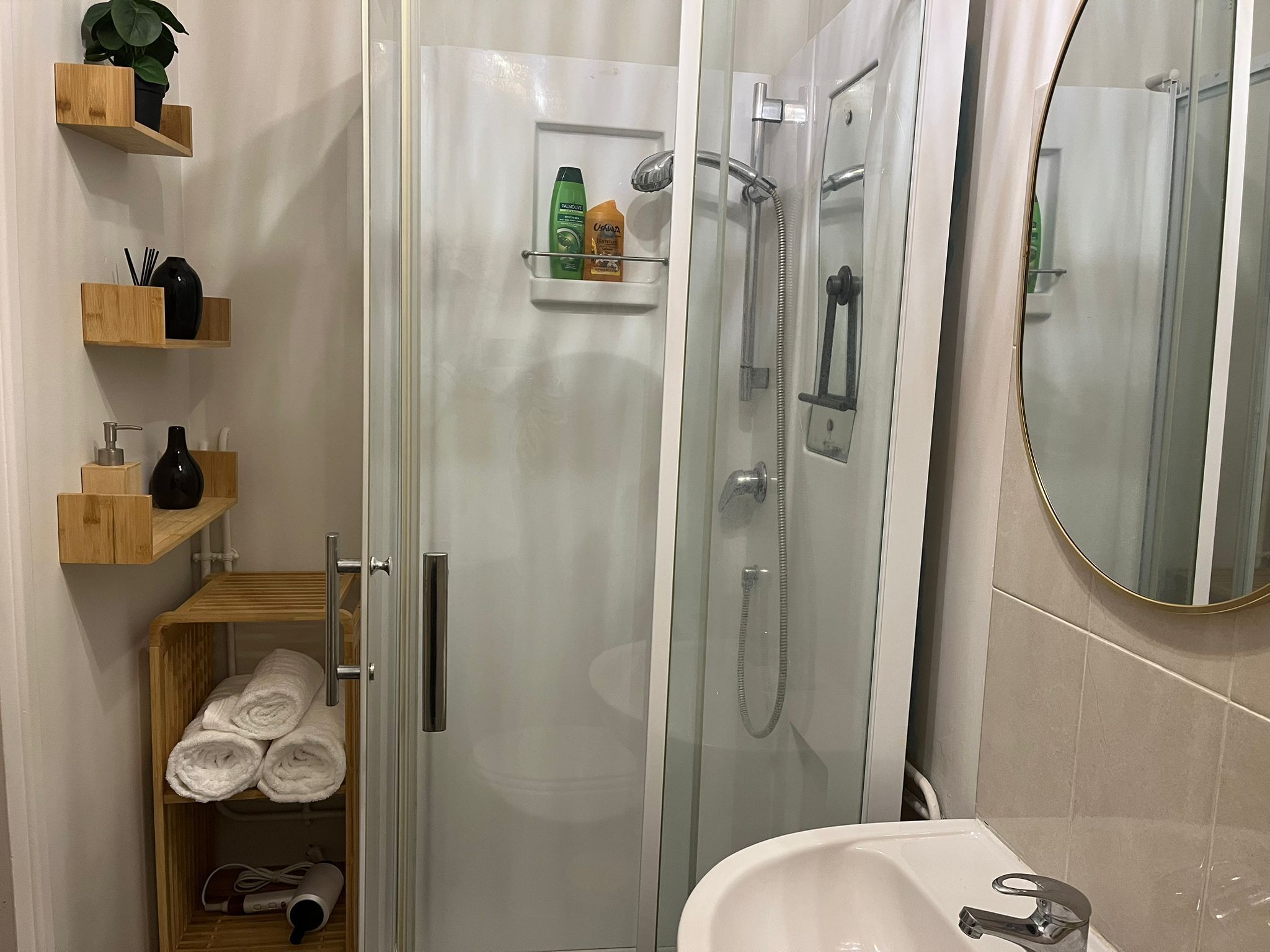 A modern bathroom with a glass-enclosed shower, a wooden shelf with toiletries, and a mirror.