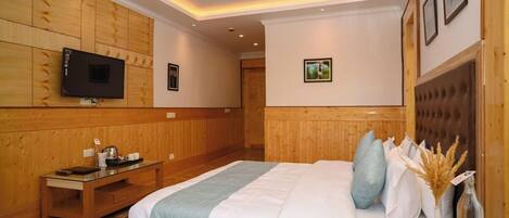 A hotel room with a wooden paneled wall, a flat-screen TV mounted above a wooden desk, a bed with a white and blue bedspread, and a small table with a vase and decorative items.