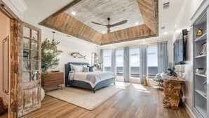 A spacious bedroom with a large bed, wooden ceiling beams, a ceiling fan, and a view of the ocean through large windows.