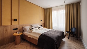 Ein modernes Hotelzimmer mit einem Holz-Kopfteil, einem großen Bett mit dunkler Tagesdecke, einem Nachttisch mit Lampe und einem Sessel.