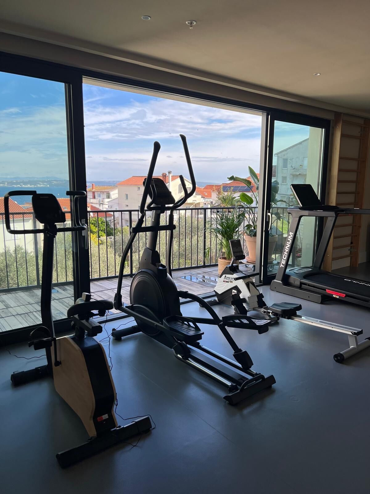 A home gym with various exercise equipment, large windows, and a view of buildings and a body of water.