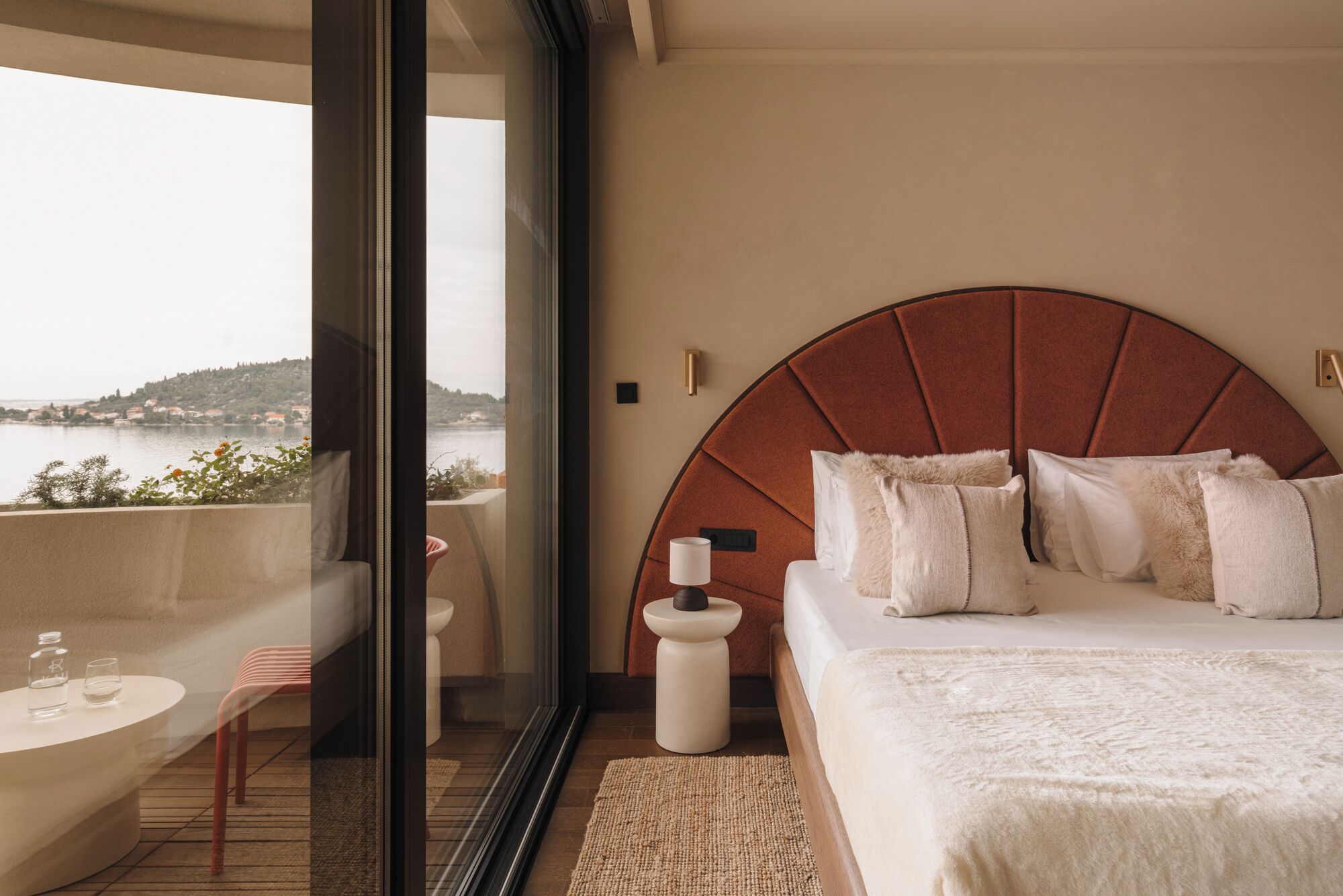 A bedroom with a large bed, a view of the sea, a glass door leading to a balcony, and a small table with a bottle and glass.