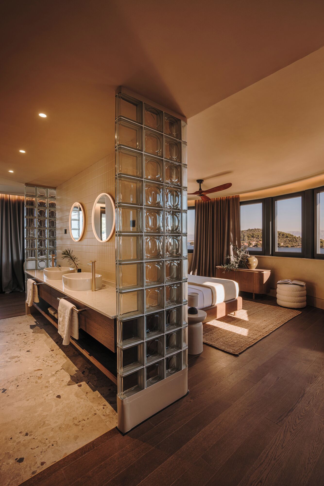 A modern bathroom with a glass partition, two sinks, a large mirror, and a view of the outdoors.