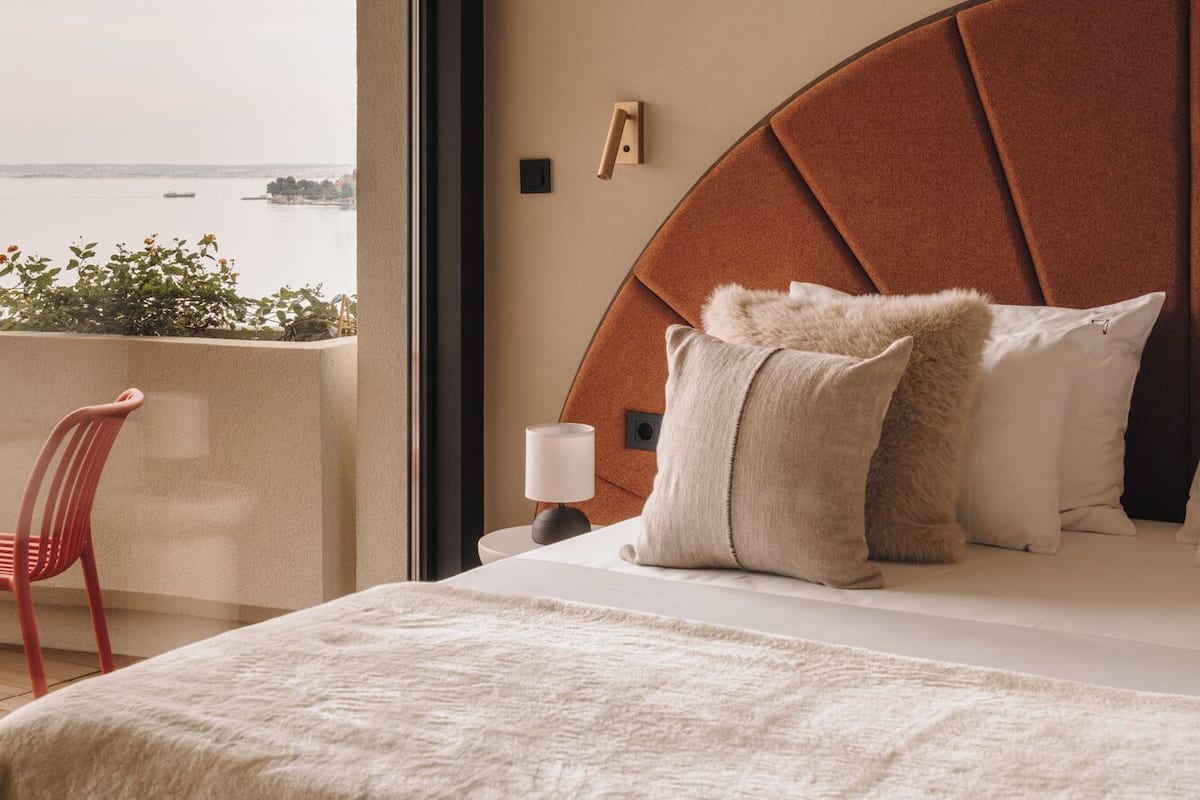 A bedroom with a large bed, a red chair, a nightstand with a lamp, and a view of the sea.