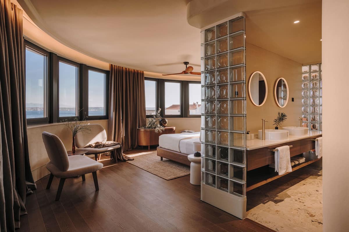 A modern hotel room with a large window, a seating area with a chair and a small table, a bed, a vanity with a mirror, and a glass block partition.