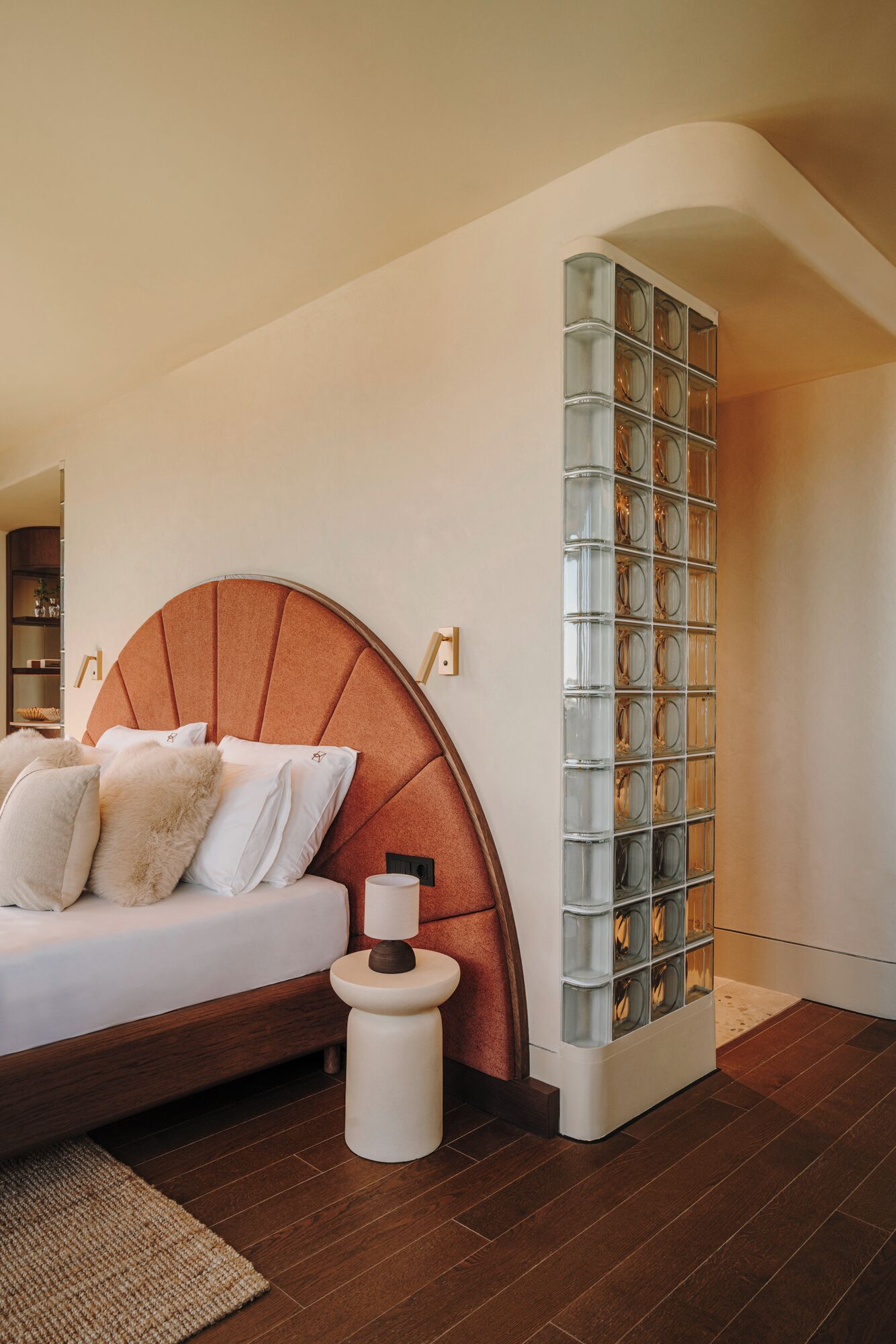 A modern bedroom with a curved headboard, a glass block wall, and a wooden floor.
