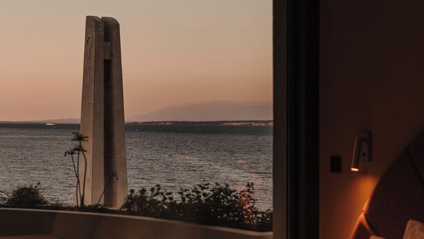 A modern hotel room with a large bed, a small table with two glasses, a chair, and a view of the sea and a tall structure at sunset. - Hotel Kali (Kali)