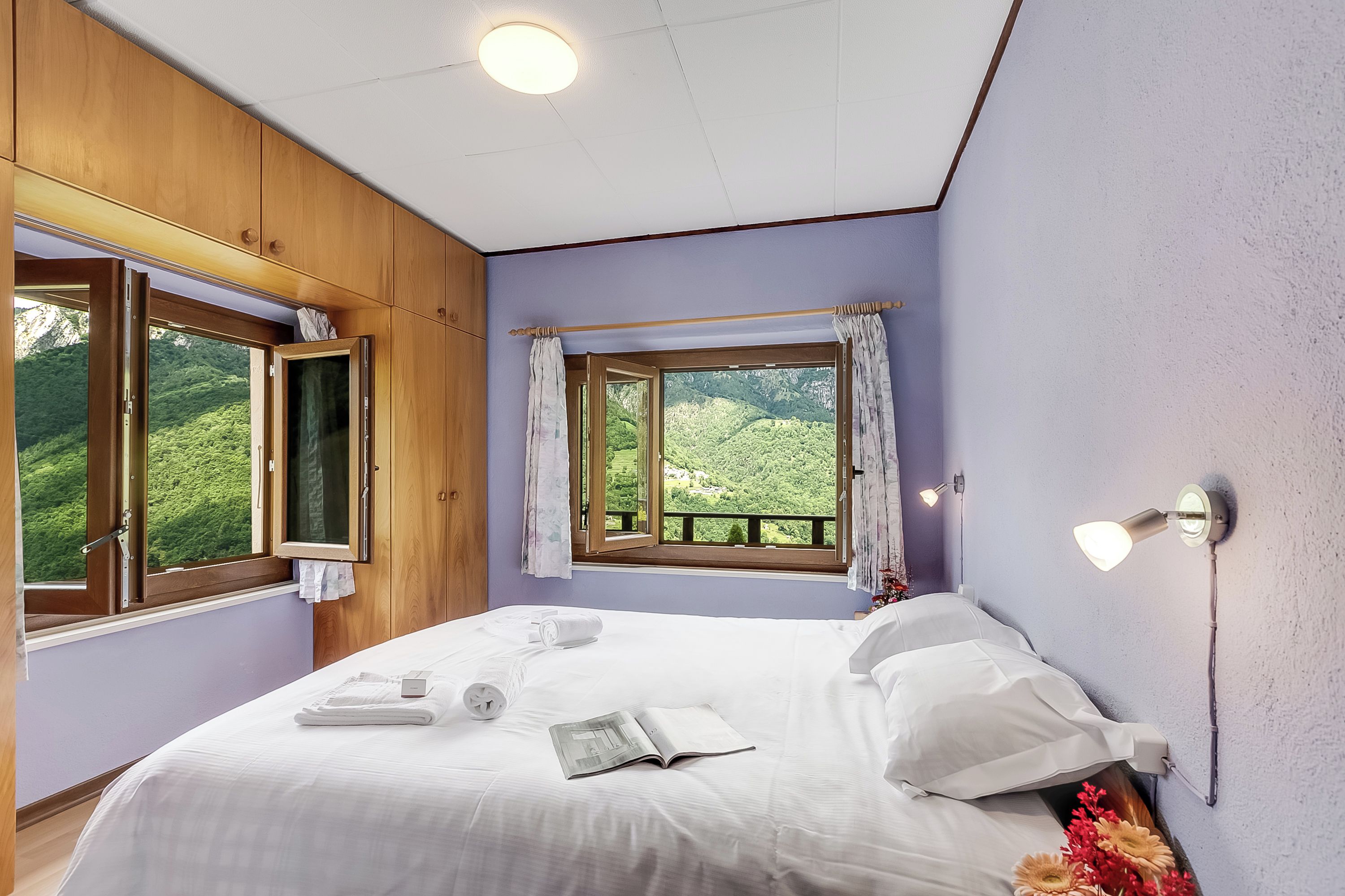 A bedroom with a bed, a window with curtains, a bedside table, and a view of greenery outside.