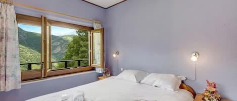 Un dormitorio con una cama grande, una ventana con vistas a colinas verdes, una mesita de noche con lámpara y una mesita auxiliar con un arreglo floral.