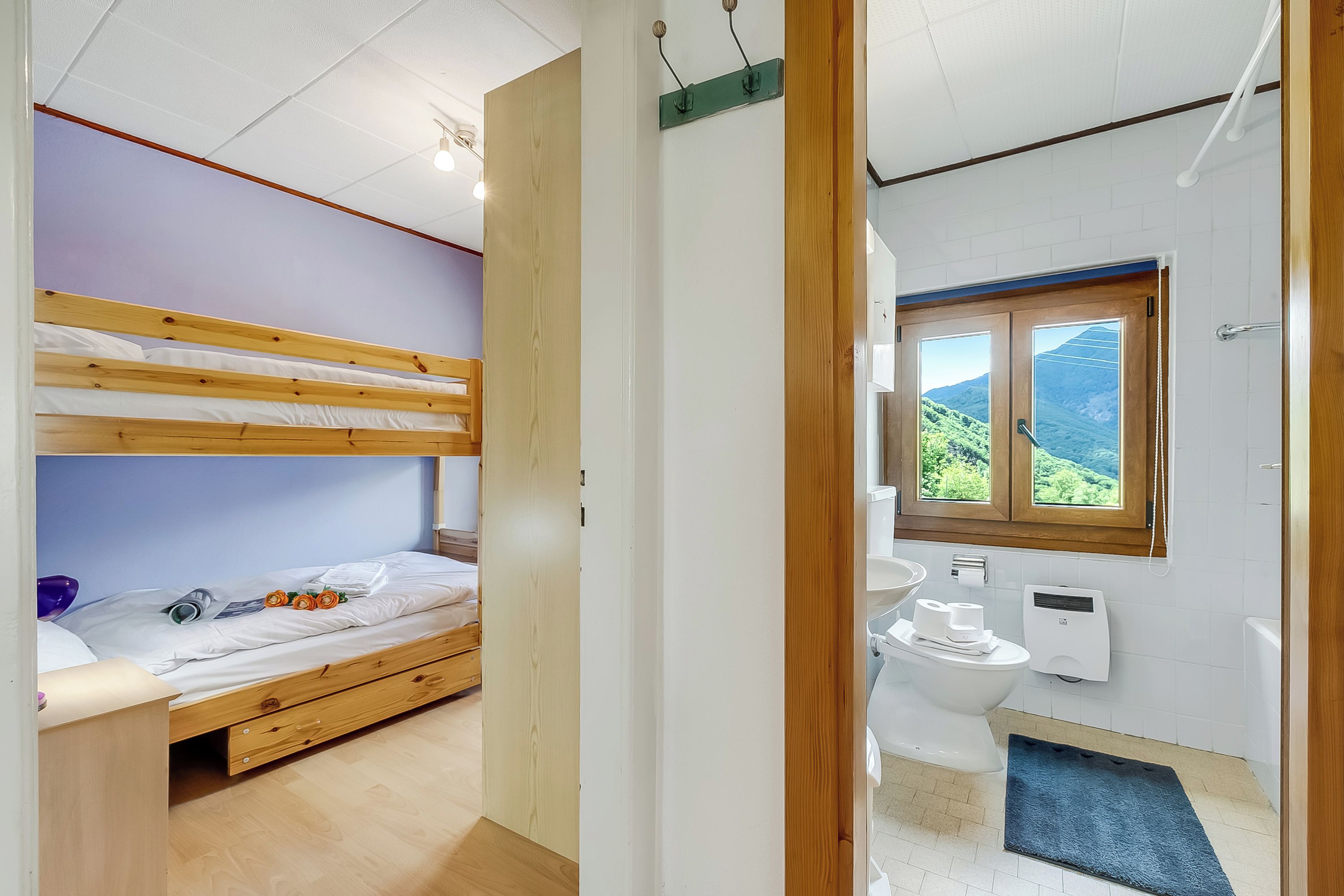 A room with a bunk bed, a wooden door, a bathroom with a toilet, a sink, and a window with a view of greenery.