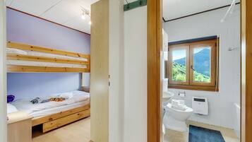 A room with a bunk bed, a wooden door, a bathroom with a toilet, a sink, and a window with a view of greenery.