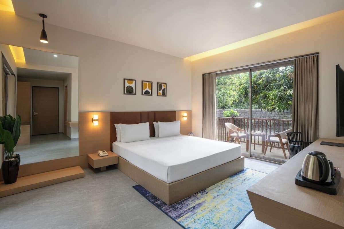 A modern hotel room with a large bed, bedside tables, a desk, and a balcony with outdoor seating.