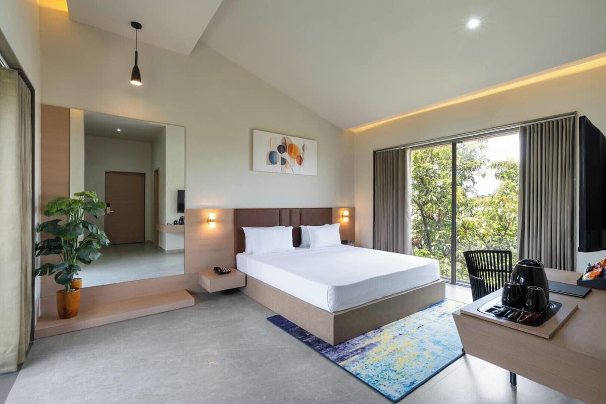 A modern hotel room with a large bed, bedside tables, a desk with a chair, a potted plant, and a view of greenery through the window.