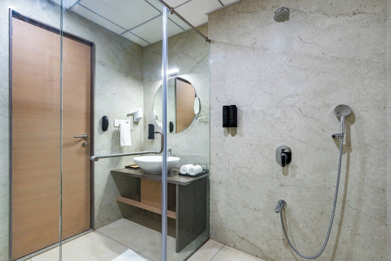 A modern bathroom with a glass shower enclosure, a wooden door, a marble wall, a white sink, and a handheld showerhead.