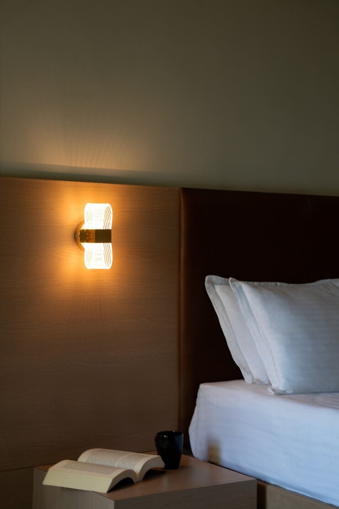 A neatly made bed with white linens, a bedside table with a book and a black mug, and a wall-mounted light fixture.