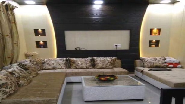 A living room with a sectional sofa, a coffee table, and a TV mounted on the wall. - Modern Apartment with Kitchen & Wi-Fi – Perfect for Work & Relaxation (Sahibzada Ajit Singh Nagar)