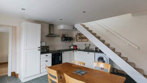 A compact kitchen with white cabinets, a dining table with chairs, a washing machine, and a staircase. - Elm House Cottage: Charming Country Cottage with Log Burner (High Hesket)