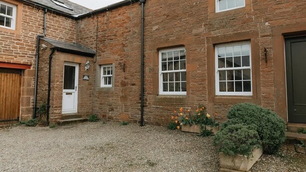 A stone building with a gravel driveway, a wooden door, and a small garden with flowers and a bush. - Elm House Cottage: Charming Country Cottage with Log Burner (High Hesket)