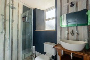 Un baño con cabina de ducha de vidrio, un lavamanos blanco y un dispensador de jabón verde, y una ventana sobre el inodoro.