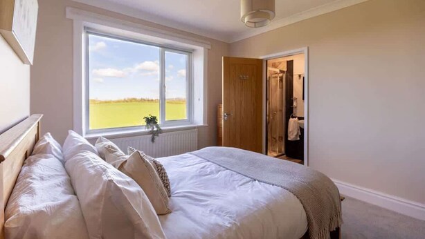 Un dormitorio con un ventanal grande, una cama con ropa de cama blanca, una lámpara colgante y una puerta de madera que da a un baño.