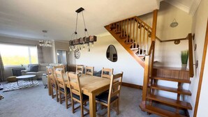 Amplia sala de estar con una mesa y sillas de madera, una escalera y un gran ventanal que proporciona luz natural.