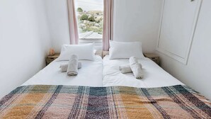 A neatly made bed with white linens and rolled towels, a colorful striped rug, and a view of greenery through the window. - Charming Ulverston Cottage with Garden & Views (Westmorland and Furness)