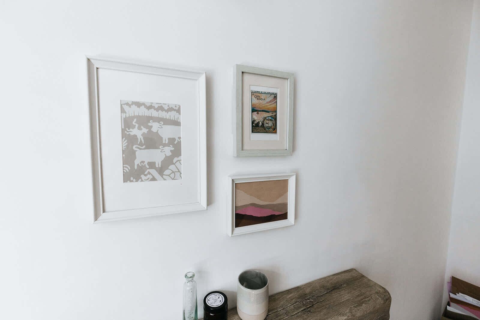 Three framed artworks on a wall, a wooden bench, and a small table with a bottle and a container.