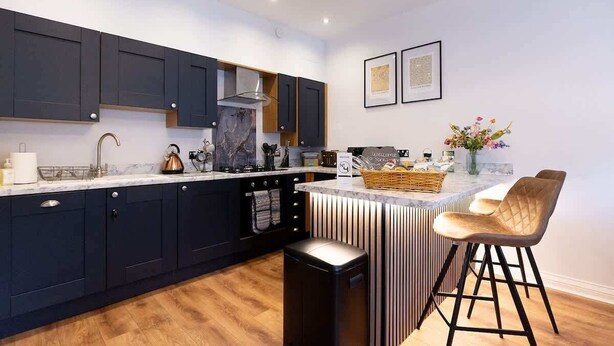 Une cuisine moderne avec des meubles de couleur sombre, un plan de travail en marbre et un îlot central doté de tabourets de bar.