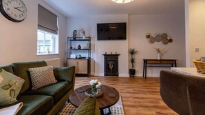 Un salon avec un canapé vert, une table basse ornée de fleurs, une cheminée, un téléviseur fixé au mur et un lustre.