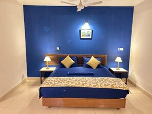 A bedroom with a blue wall, a wooden bed frame, two bedside tables with lamps, and a ceiling fan. - Kamat Holiday Home (Calangute)