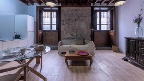 A spacious living area with a glass dining table, a sofa, and a wooden coffee table. There are two windows with curtains, a stone wall, and a dark wooden cabinet.