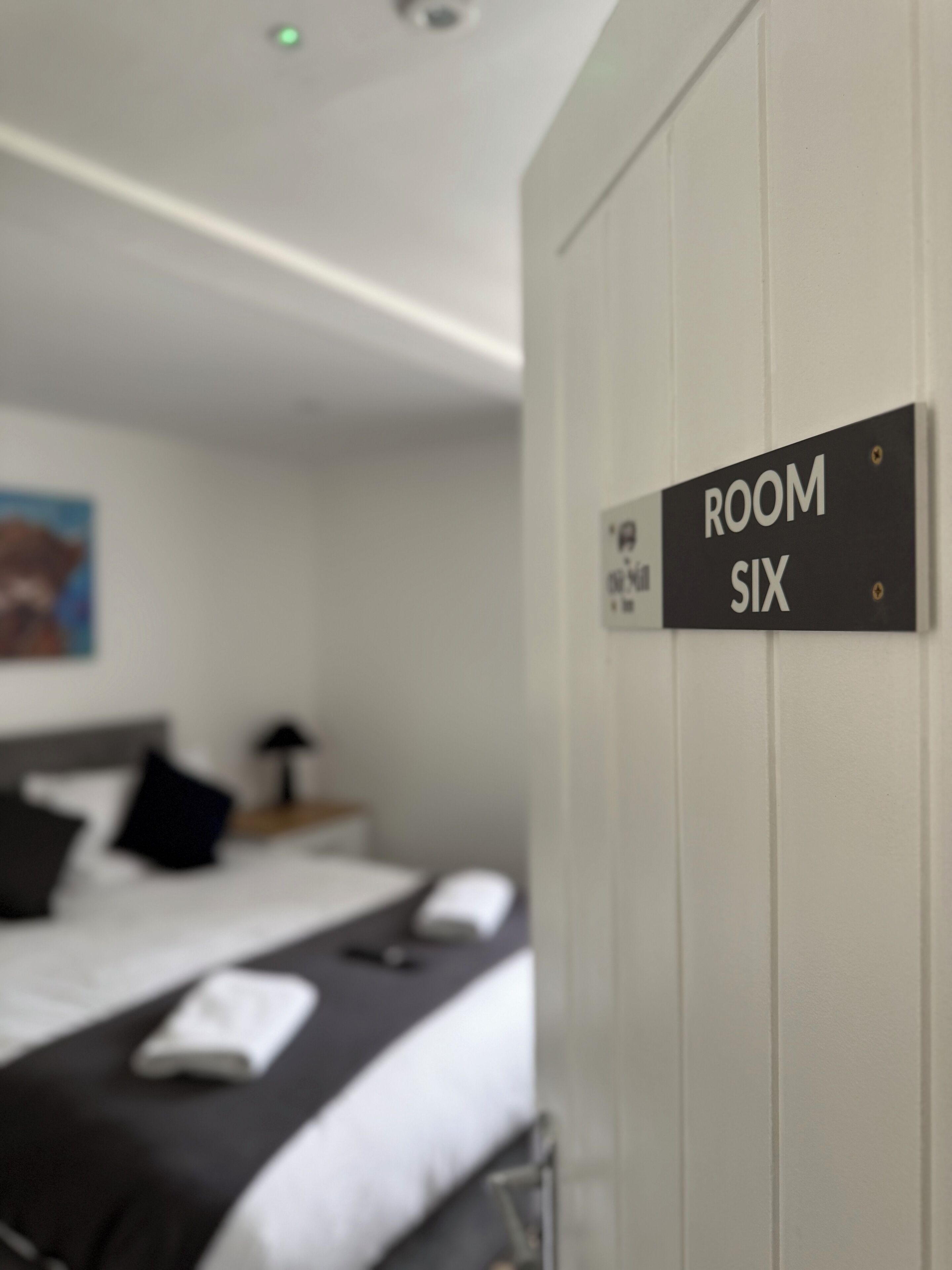 Una señal que indica 'ROOM SIX' en una habitación de hotel con una cama, almohadas y una mesita de noche.