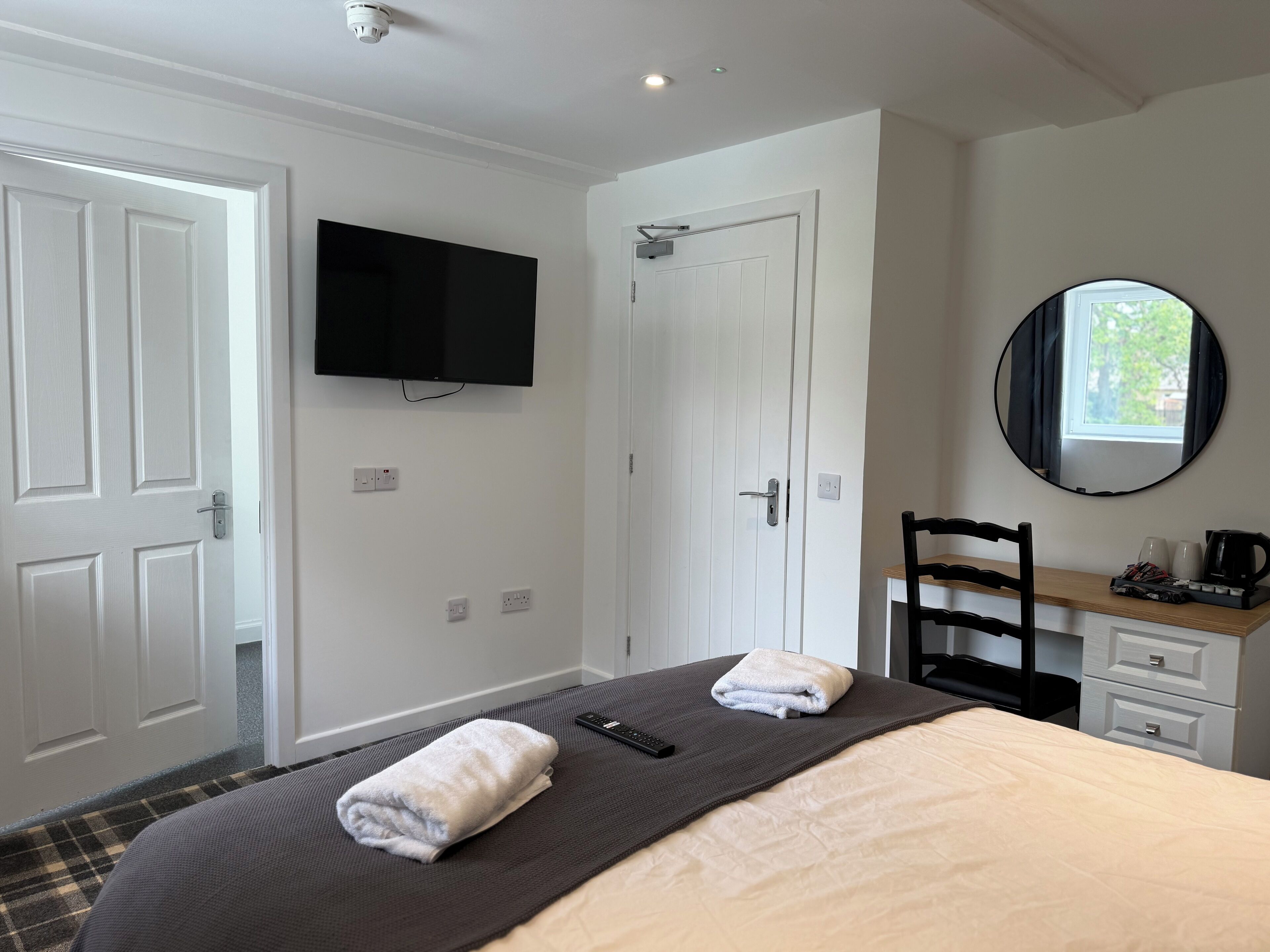 A hotel room with a bed, a television mounted on the wall, a mirror, a desk with a chair, and a window.