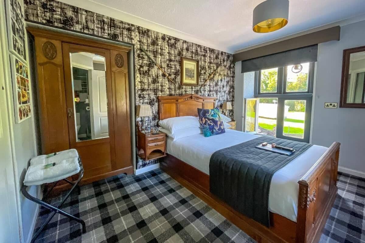 A bedroom with a checkered floor, a wooden bed with a headboard, a nightstand, a mirror, and a window with a view of greenery.