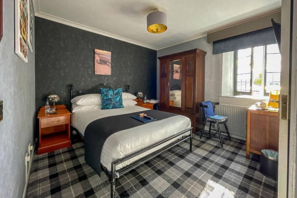 A bedroom with a checkered floor, a bed with a black metal frame, a wooden nightstand, a desk, and a chair.