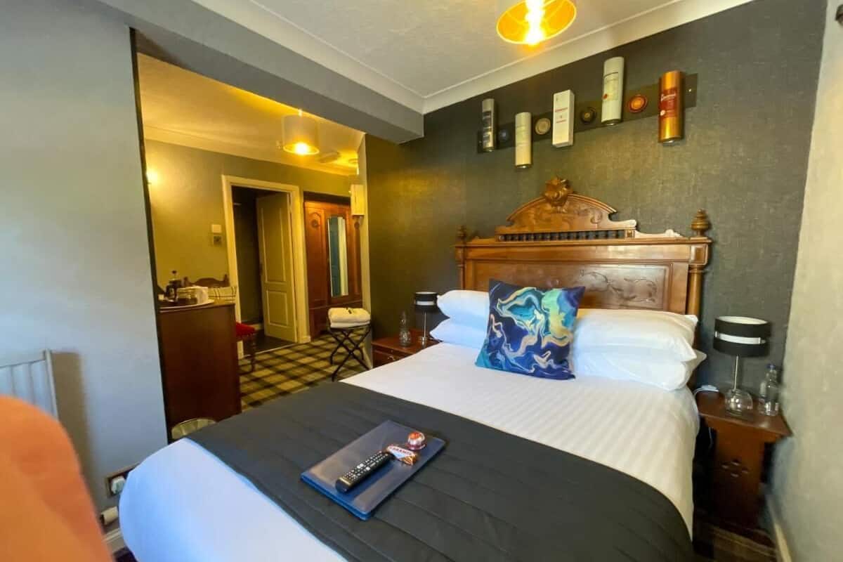 A hotel room with a large bed, a wooden headboard, a nightstand with a lamp, and a wall-mounted shelf with toiletries.