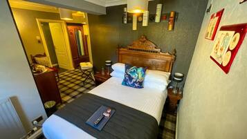 A hotel room with a large bed, a wooden headboard, a checkered floor, a desk with a chair, and a lamp.