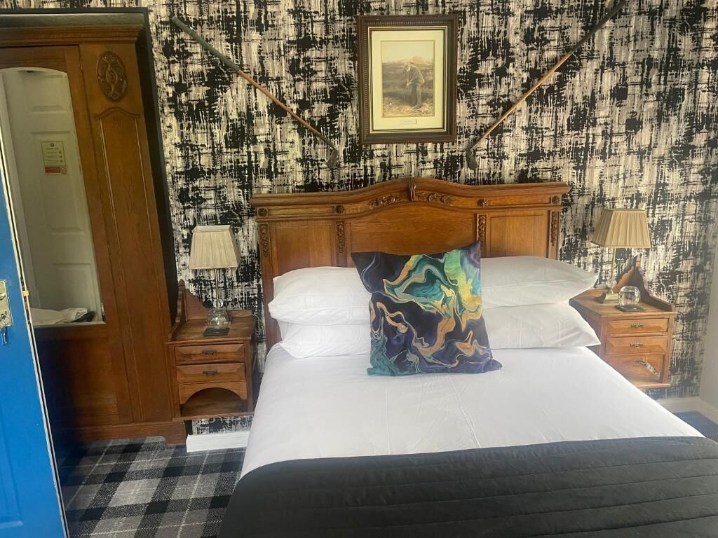 A hotel room with a bed, a wooden wardrobe, a nightstand with a lamp, and a wall with a framed picture.