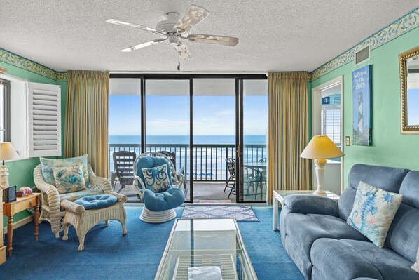 A room with a view of the ocean, featuring wicker furniture, a blue sofa, and a ceiling fan.