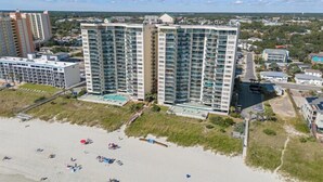 A high-rise hotel with multiple balconies, a swimming pool, and a beach area with umbrellas.
