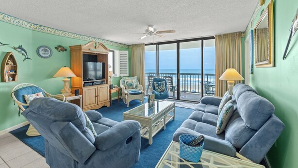A spacious living room with a large window offering a view of the ocean, a ceiling fan, and a television on a wooden cabinet.