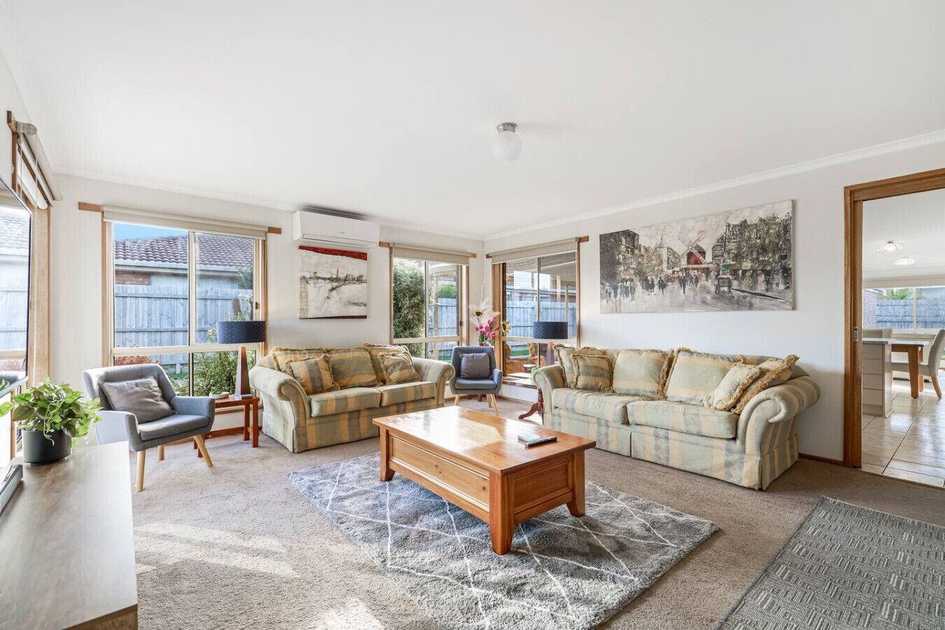 A spacious living room with two sofas, a coffee table, and a TV. There are large windows and a painting on the wall.