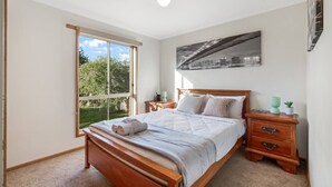 Ein Schlafzimmer mit großem Fenster, einem Bett mit Holz-Kopfteil, einem Nachttisch und einem an der Wand hängenden Bild.