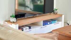 Ein Wohnzimmer mit Flachbildfernseher auf einem Holzboden, einer kleinen Pflanze und einem Bücherregal mit Büchern und Spielen.