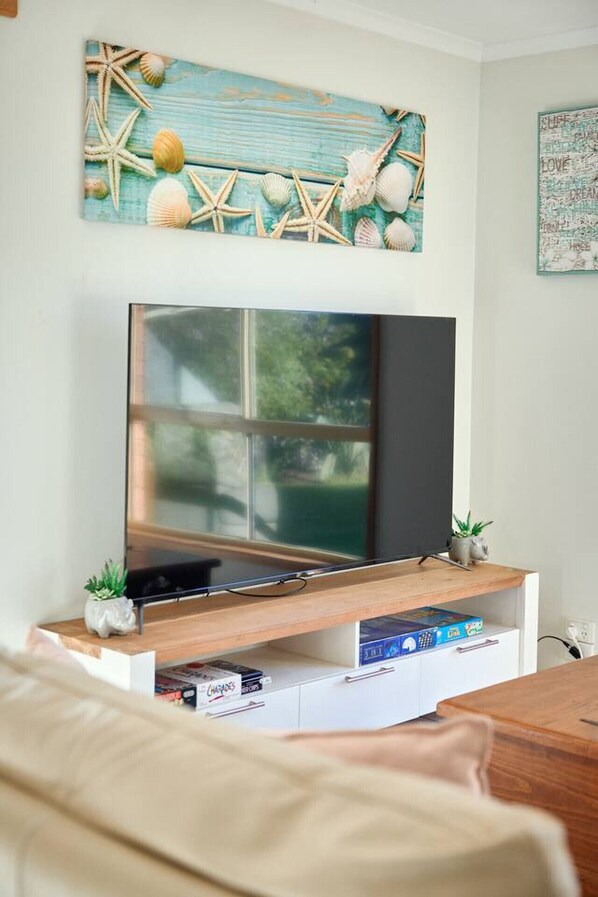 A living room with a flat-screen TV on a wooden stand, a small plant, and a bookshelf with books and games.