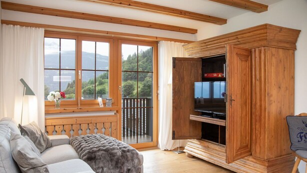 A cozy living room with a sofa, a wooden cabinet with a television, and a balcony with a view of the mountains. - Beautiful apartment for 5 people with pool, WIFI, TV, balcony and panoramic view (Falera)