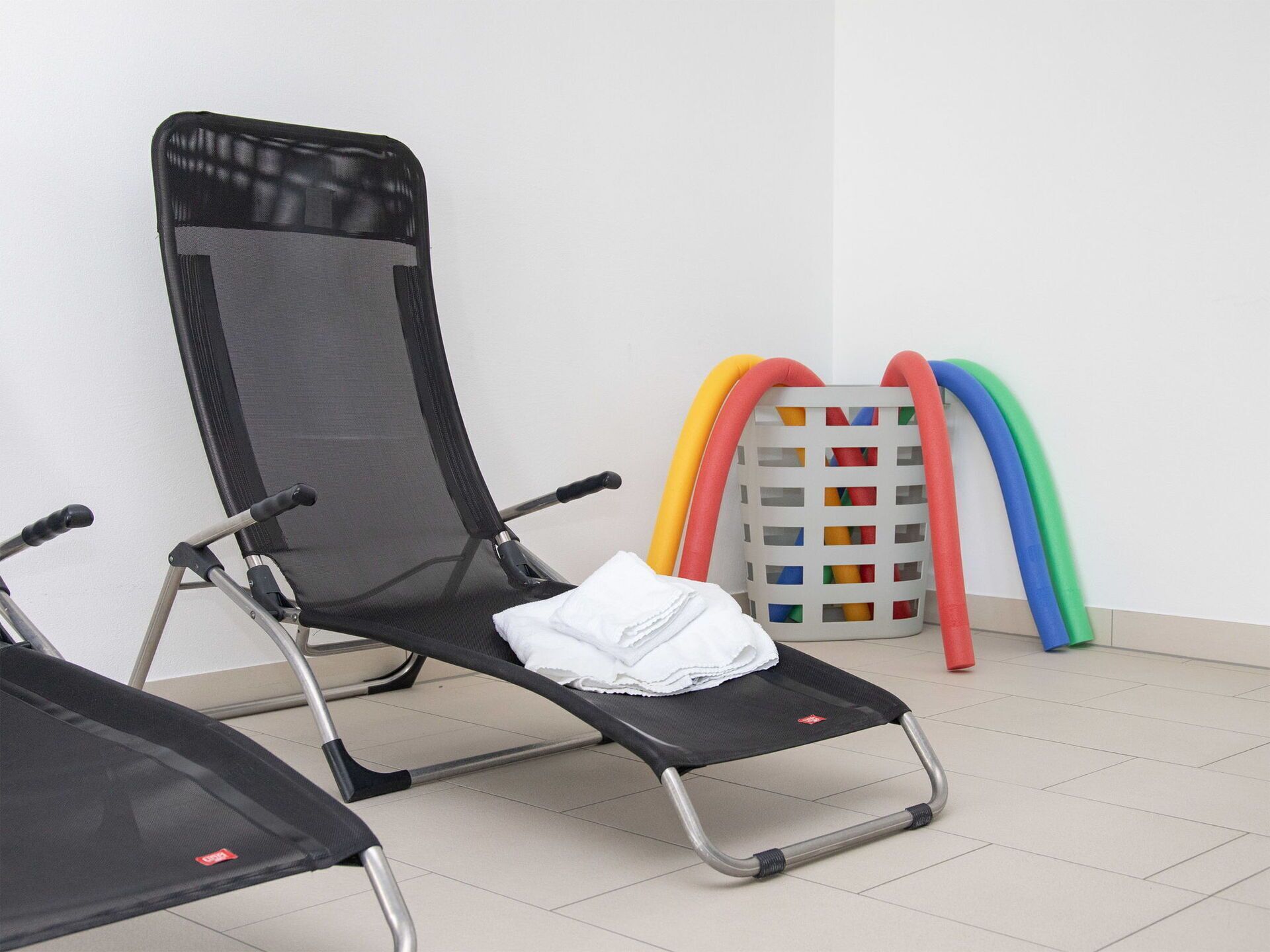 Una habitación con una silla reclinable negra, una toalla blanca sobre ella, y equipo de ejercicio de colores contra la pared.