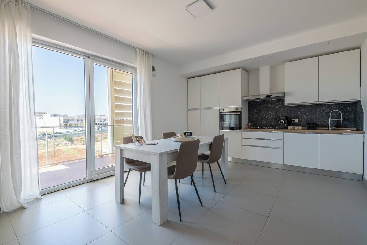 Una cocina moderna con armarios blancos, una mesa de comedor con sillas y un amplio ventanal corredero que da a un balcón.