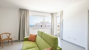 A modern living room with a green sofa, a wooden chair, and large windows offering a view of buildings and the sea.