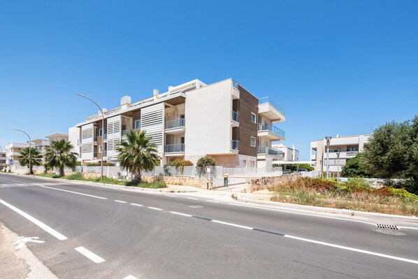 A modern multi-story residential building with balconies, a paved road in front, and palm trees. - White Coast Relax & Stay (Polignano a Mare)